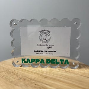 May include: A clear acrylic magnetic photo frame with a scalloped edge. The frame has the text "KAPPA DELTA" in green at the bottom. The frame holds 3x5 or 4x6 photos and has the text "thehennhouse by jenn" on the front.
