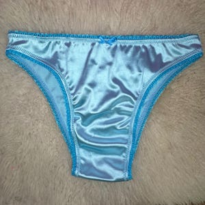 Könnte beinhalten: Hellblaue Satin-Unterwäsche mit hellblauer Spitzenborte. Eine kleine, hellblaue Schleife befindet sich vorne. Die Unterwäsche besteht aus einem glänzenden, glatten Stoff. Ein Wäschestück.