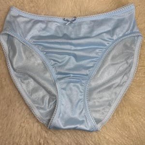 Puede incluir: Braga de corte alto en color azul claro con un acabado satinado suave y un delicado borde de encaje blanco a lo largo de la cintura y las aberturas de las piernas. Un pequeño lazo de cinta azul claro a juego está centrado en la cintura.