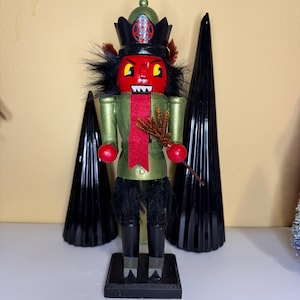 Op de afbeelding: Een decoratieve notenkraker met een rood gezicht, zwart haar en een groene jas. De notenkraker heeft een zwarte hoed met een rood embleem en houdt een kleine bezem vast. Twee zwarte kegelvormige decoraties staan achter de notenkraker.