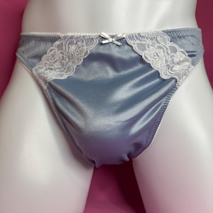 Può includere: Uno slip in raso blu ardesia con finiture in pizzo floreale bianco sui lati superiori e un piccolo fiocco in raso bianco centrato sulla vita. Il capo presenta una finitura liscia e lucente e un taglio classico con bordi elastici bianchi.