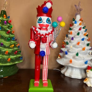 Puede incluir: Un cascanueces de madera a rayas rojas y blancas vestido de payaso con un sombrero rojo y blanco, un pompón blanco en el sombrero y un pompón rojo en el cuello. El cascanueces sostiene un palo plateado con tres globos de colores. El cascanueces tiene una cara blanca con ojos negros pintados, una boca roja y una nariz roja. El cascanueces tiene un cuerpo a rayas rojas y blancas con un pompón blanco en el pecho. El cascanueces está de pie sobre una base verde. El cascanueces tiene un botón rojo en el pecho que dice "I'm the Pogo".