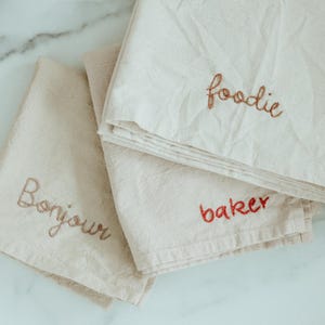 May include: Three white cotton tea towels with embroidered text. The top towel says "foodie", the middle towel says "baker", and the bottom towel says "bonjour".