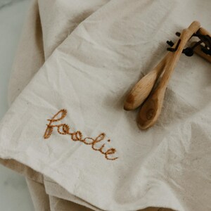 May include: A white linen tea towel with the word "foodie" embroidered in brown thread. Two wooden spoons are lying on top of the towel.