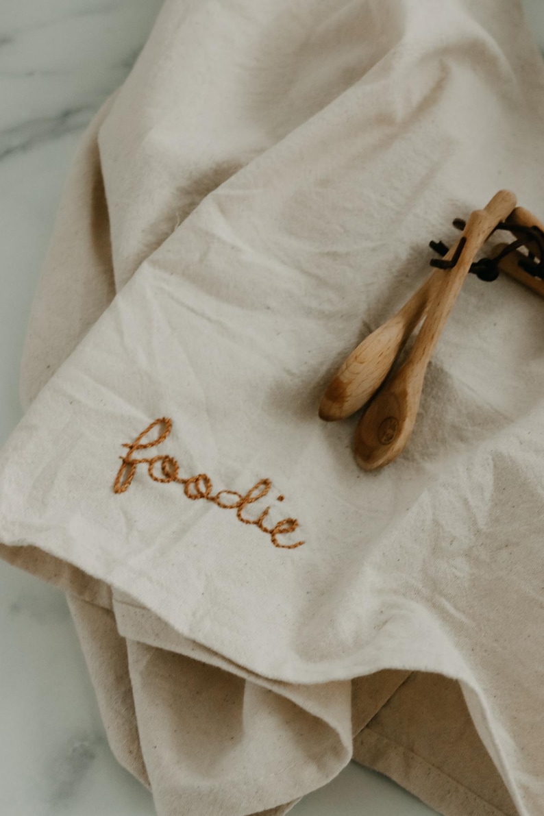 May include: A white linen tea towel with the word "foodie" embroidered in brown thread. Two wooden spoons are lying on top of the towel.
