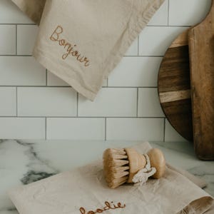 May include: Two beige kitchen towels with embroidered text. The top towel says "Bonjour" and the bottom towel says "foodie". A wooden brush is on top of the folded towel.