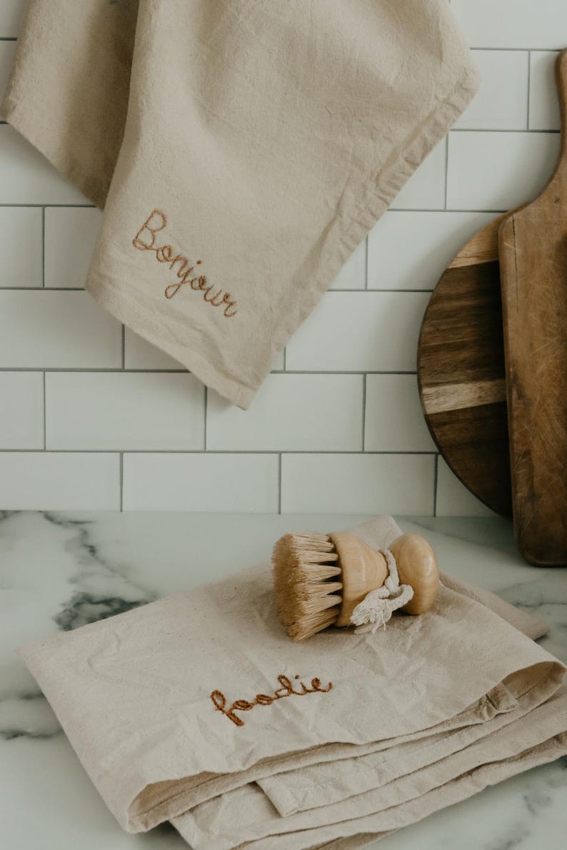 May include: Two beige kitchen towels with embroidered text. The top towel says "Bonjour" and the bottom towel says "foodie". A wooden brush is on top of the folded towel.