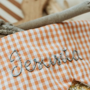 May include: A sleep mask in orange and white gingham, embroidered with the name "Jesuta" in grey thread. The image also shows a grey stuffed animal and a brown fabric tube.