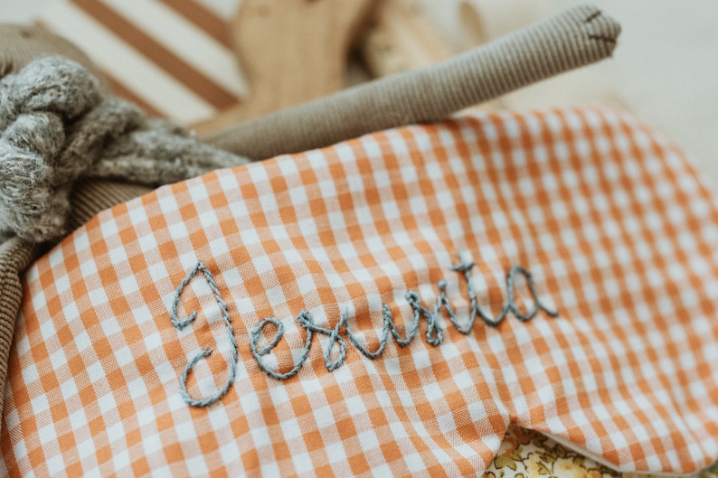 May include: A sleep mask in orange and white gingham, embroidered with the name "Jesuta" in grey thread. The image also shows a grey stuffed animal and a brown fabric tube.