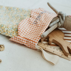 May include: A fabric stocking with a floral pattern and a gingham top, filled with a stuffed bunny, wooden toy, ruler, and other small items. The stocking has the word "sweetie" embroidered on it. Gold bells and dried flowers are scattered around.