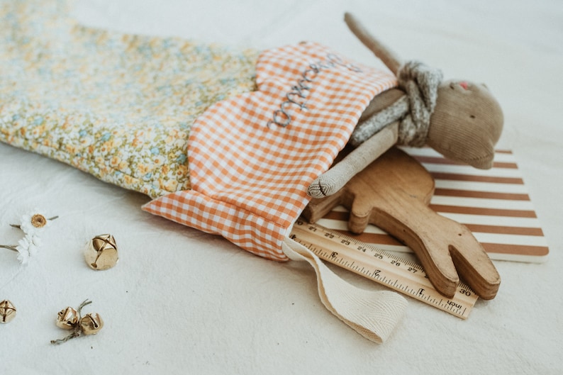 May include: A fabric stocking with a floral pattern and a gingham top, filled with a stuffed bunny, wooden toy, ruler, and other small items. The stocking has the word "sweetie" embroidered on it. Gold bells and dried flowers are scattered around.
