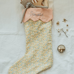 May include: A floral Christmas stocking with a scalloped orange gingham cuff embroidered with the name "Jessica". The stocking is filled with a stuffed animal, wooden toy, and other small gifts. Gold bells and a sprig of white flowers are scattered nearby.