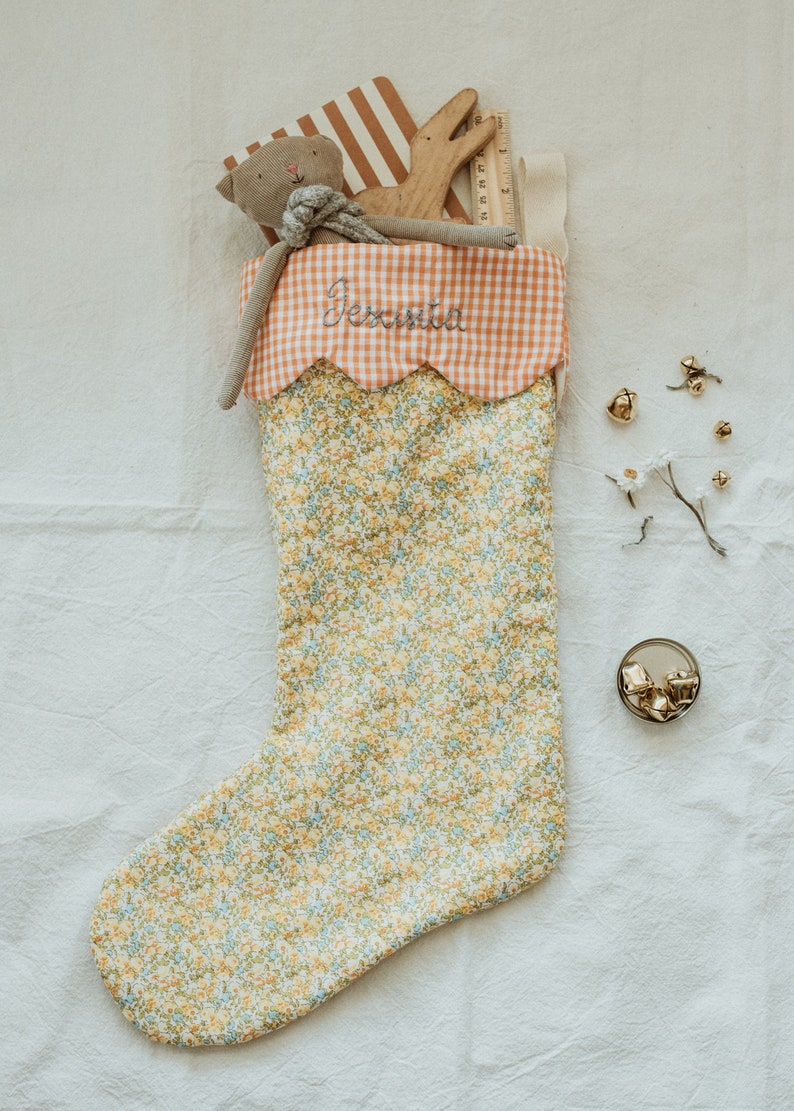 May include: A floral Christmas stocking with a scalloped orange gingham cuff embroidered with the name "Jessica". The stocking is filled with a stuffed animal, wooden toy, and other small gifts. Gold bells and a sprig of white flowers are scattered nearby.