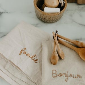 May include: Two beige kitchen towels with embroidered text. One towel says "foodie" and the other says "Bonjour". Wooden spoons are on top of the towel with the French greeting.