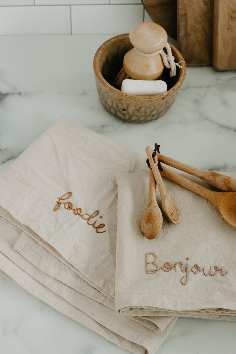May include: Two beige kitchen towels with embroidered text. One towel says "foodie" and the other says "Bonjour". Wooden spoons are on top of the towel with the French greeting.