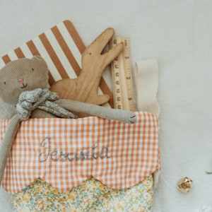 May include: A stocking filled with a stuffed cat, wooden alligator, and ruler. The stocking features an orange and white gingham pattern and the name "Jesinta" embroidered on it. The stocking is trimmed with a floral pattern.