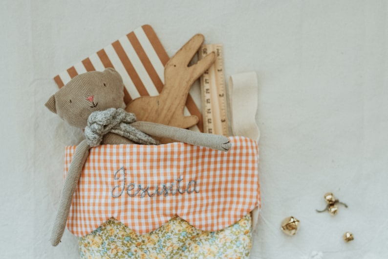 May include: A stocking filled with a stuffed cat, wooden alligator, and ruler. The stocking features an orange and white gingham pattern and the name "Jesinta" embroidered on it. The stocking is trimmed with a floral pattern.