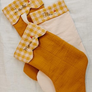May include: Three Christmas stockings in mustard yellow and cream. The top of each stocking has a scalloped edge with a yellow and white gingham pattern. Each stocking is embroidered with a name and a small mushroom.