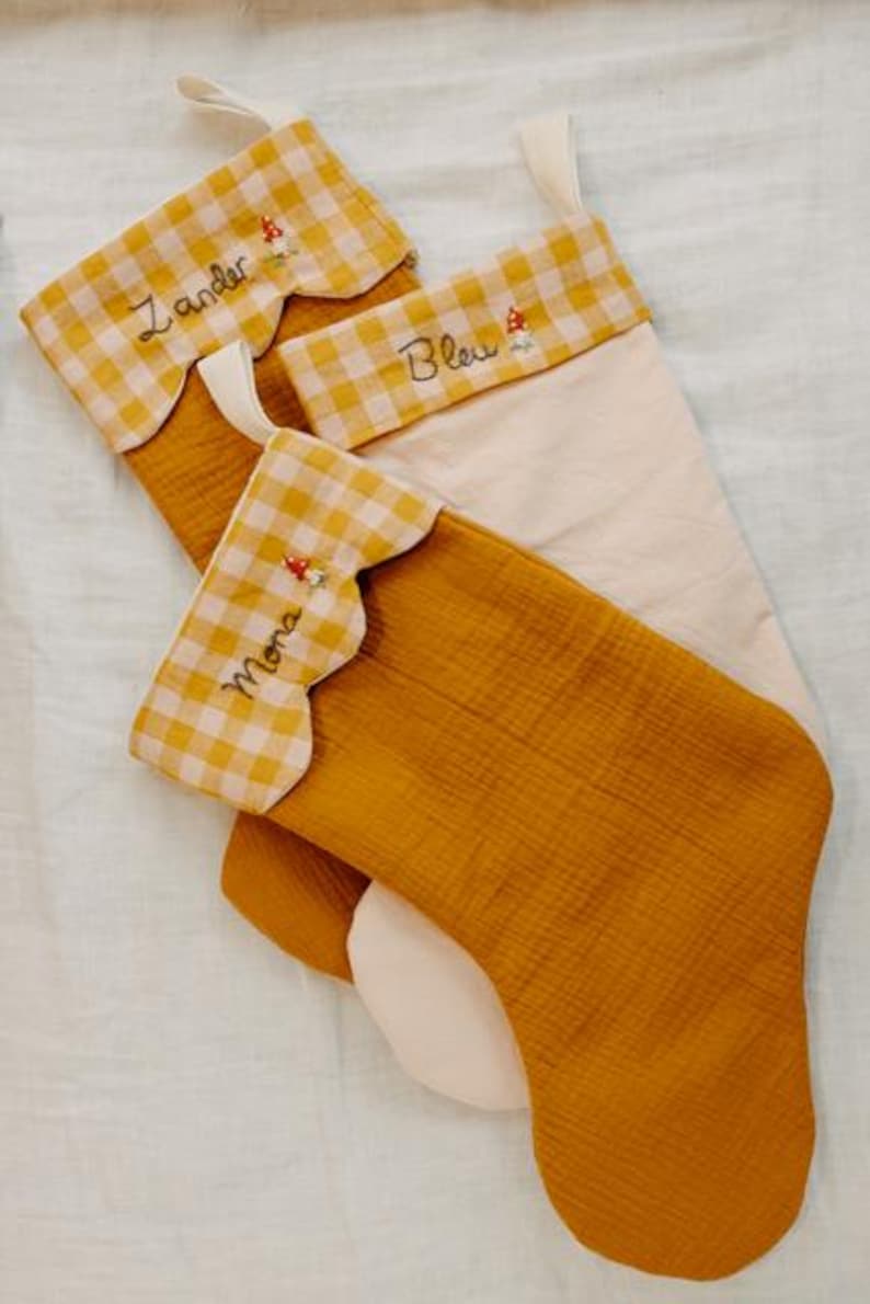 May include: Three Christmas stockings in mustard yellow and cream. The top of each stocking has a scalloped edge with a yellow and white gingham pattern. Each stocking is embroidered with a name and a small mushroom.