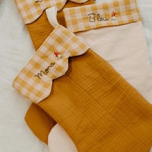 May include: Three Christmas stockings in mustard yellow and cream. The top of each stocking has a scalloped edge with a yellow and white gingham pattern. Each stocking is personalised with a name embroidered in cursive and a small mushroom.