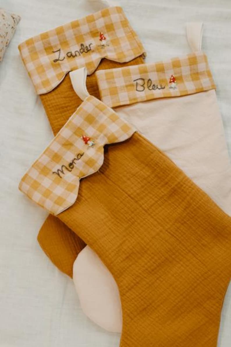 May include: Three Christmas stockings in mustard yellow and cream. The top of each stocking has a scalloped edge with a yellow and white gingham pattern. Each stocking is personalised with a name embroidered in cursive and a small mushroom.