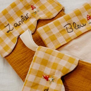 May include: Close-up of three Christmas stockings. The stockings are made of mustard-coloured fabric with a yellow and white gingham pattern on the cuff. Each stocking has a name embroidered on it, along with a small mushroom design.