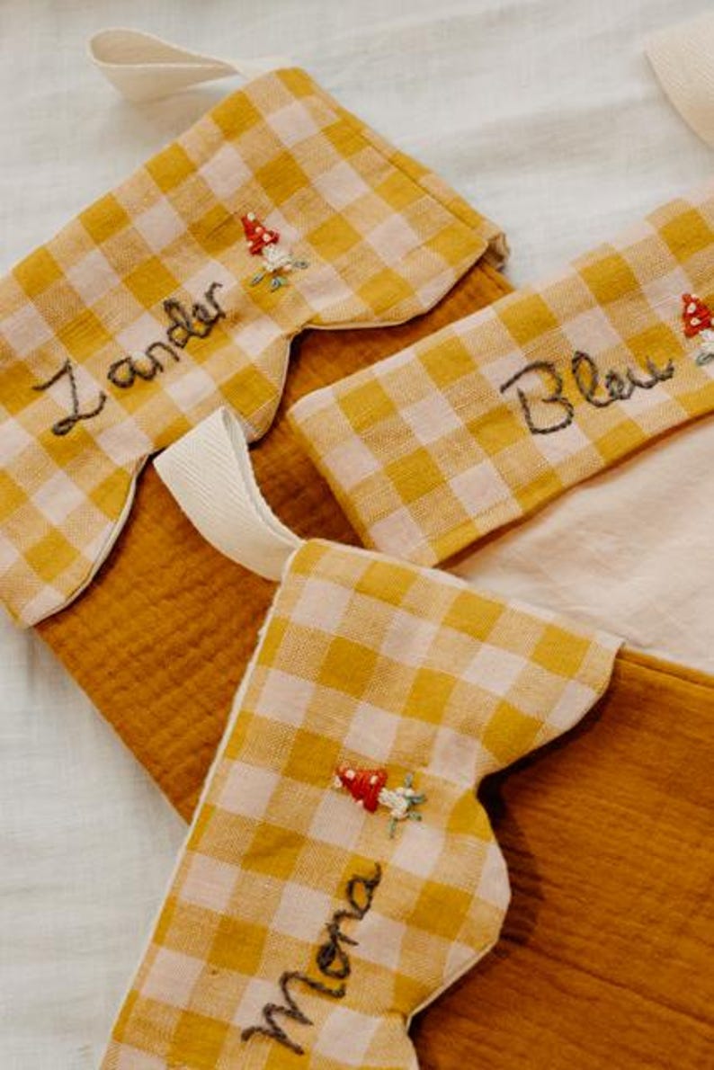 May include: Close-up of three Christmas stockings. The stockings are made of mustard-coloured fabric with a yellow and white gingham pattern on the cuff. Each stocking has a name embroidered on it, along with a small mushroom design.