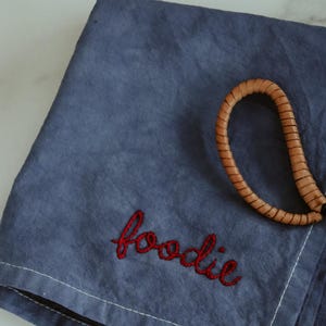 May include: A blue linen kitchen towel with the word "foodie" embroidered in red thread. A brown leather strap is on top of the towel.