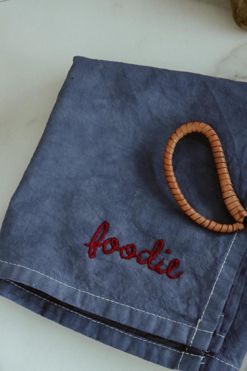 May include: A blue linen kitchen towel with the word "foodie" embroidered in red thread. A brown leather strap is on top of the towel.