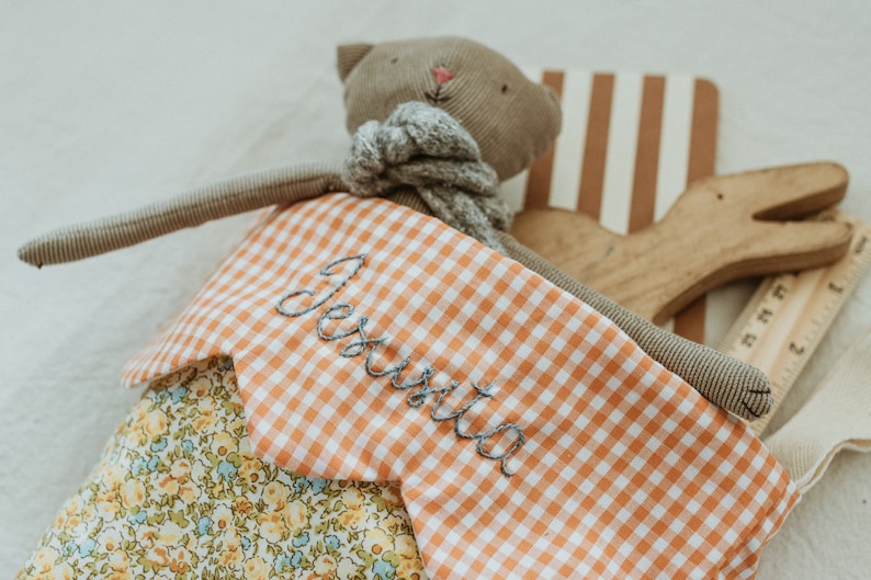 May include: A handmade stuffed cat toy with long arms and a grey scarf. The cat wears a dress with a floral pattern and an orange and white gingham bib embroidered with the name "Jes&uacute;s". A wooden toy and ruler are also visible.