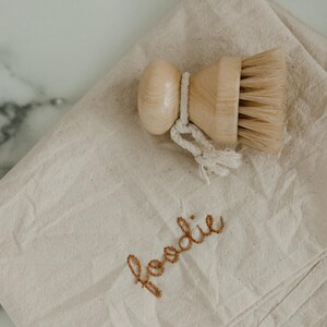 May include: A beige canvas dishcloth with the word "foodie" embroidered in brown thread. A wooden brush with natural bristles is on top of the cloth.