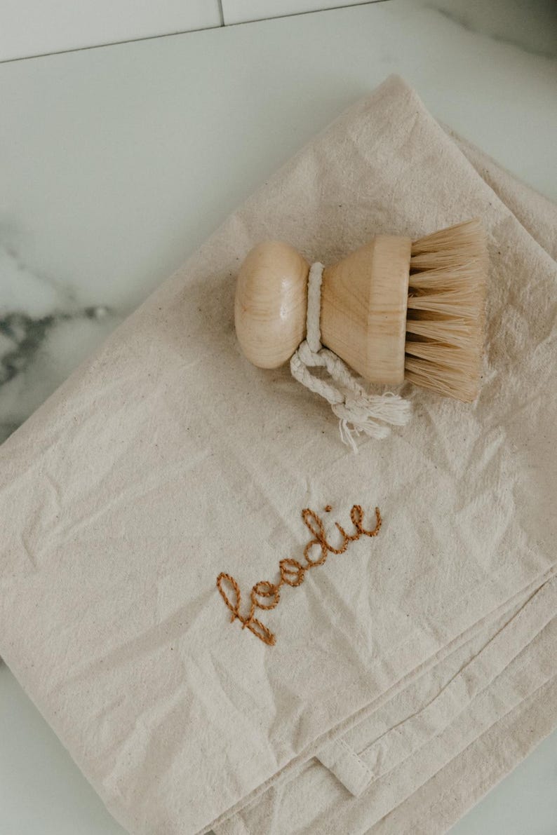 May include: A beige canvas dishcloth with the word "foodie" embroidered in brown thread. A wooden brush with natural bristles is on top of the cloth.