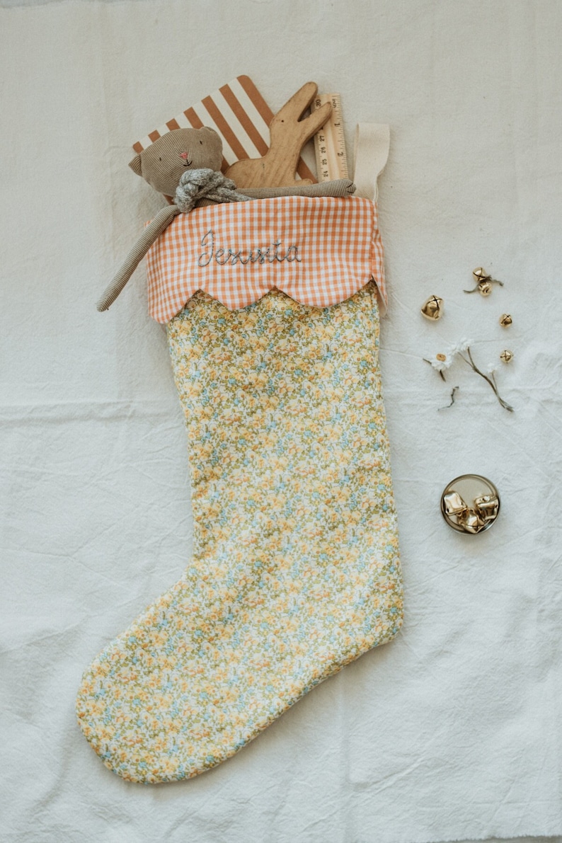 May include: A floral Christmas stocking with a gingham cuff embroidered with the name "Jessica". The stocking is filled with a stuffed animal, wooden toy, and other small gifts. Gold bells and decorative sprigs are scattered around.