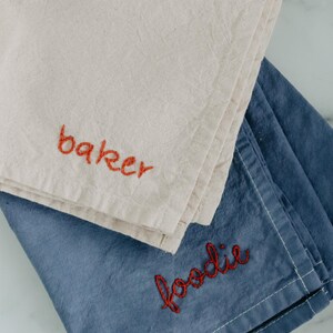 May include: Two kitchen towels, one beige and one blue, with red embroidery. The beige towel says "baker" and the blue towel says "foodie".