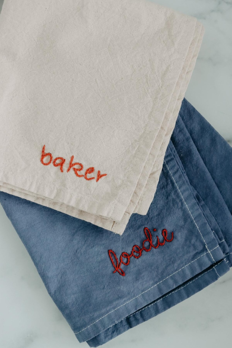 May include: Two kitchen towels, one beige and one blue, with red embroidery. The beige towel says "baker" and the blue towel says "foodie".