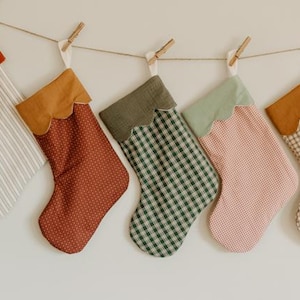 May include: Five festive Christmas stockings suspended from a string. Each stocking features a unique patterned fabric, with scalloped tops in shades of red, brown, green, and tan. The fabrics include stripes, polka dots, and gingham patterns.