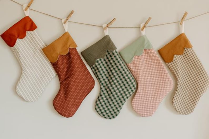 May include: Five festive Christmas stockings suspended from a string. Each stocking features a unique patterned fabric, with scalloped tops in shades of red, brown, green, and tan. The fabrics include stripes, polka dots, and gingham patterns.