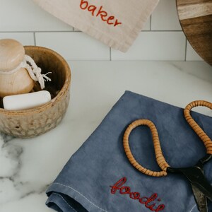 May include: Two kitchen towels with embroidered text. One towel is beige with the word "baker" embroidered in red. The other towel is blue with the word "foodie" embroidered in red. A pair of scissors is on top of the blue towel.