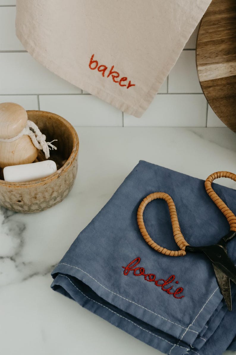 May include: Two kitchen towels with embroidered text. One towel is beige with the word "baker" embroidered in red. The other towel is blue with the word "foodie" embroidered in red. A pair of scissors is on top of the blue towel.