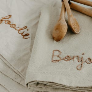 May include: Two white linen kitchen towels with embroidered text. One towel says "foodie" and the other says "Bonjour".