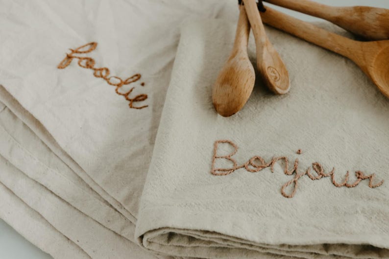 May include: Two white linen kitchen towels with embroidered text. One towel says "foodie" and the other says "Bonjour".