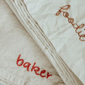 May include: Two white fabric tea towels with embroidered names. The top towel has the name "Freddie" embroidered in brown thread. The bottom towel has the name "baker" embroidered in red thread.