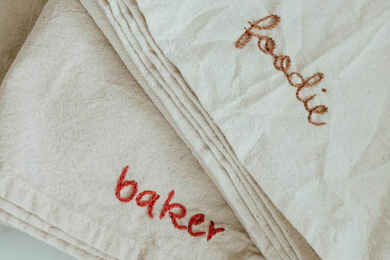 May include: Two white fabric tea towels with embroidered names. The top towel has the name "Freddie" embroidered in brown thread. The bottom towel has the name "baker" embroidered in red thread.