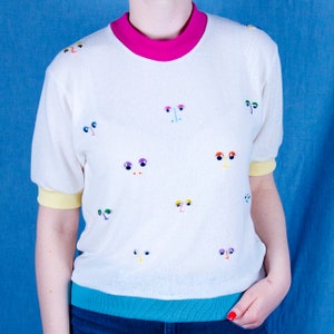 May include: White short-sleeved shirt with a pink collar and colourful embroidered eyes on the front.