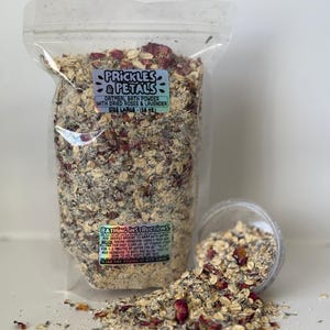 May include: A clear plastic bag filled with a mixture of oatmeal, dried rose petals, and lavender. The bag has a label that reads "Prickles & Petals Oatmeal Bath Powder with Dried Roses & Lavender Size Large (16 oz)". There is a small clear plastic container with a scoop of the bath powder on a white surface.