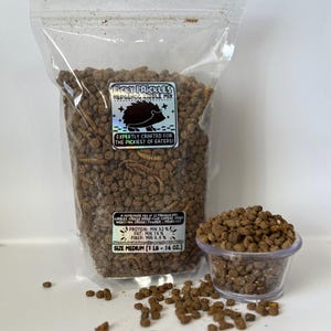 May include: A clear plastic bag filled with brown hedgehog kibble mix. The bag has a label that says "Picky Prickles Hedgehog Kibble Mix" and "Expertly Crafted for the Pickiest of Eaters!" The label also lists the nutritional information: Protein: Min 32%, Fat: Min 14%, Fiber: Min 5.5%. The bag is labeled "Size Medium (1 lb - 16 oz.)". A small clear plastic cup filled with the kibble mix is sitting on a white surface.