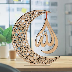 Könnte beinhalten: Ein Holz-Halbmond mit ausgeschnittenen Sternen, an dem eine Holz-Tränenform mit dem arabischen Wort "Allah" eingeschrieben hängt. Das Stück steht auf einem Holzsockel.