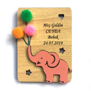 May include: A wooden plaque with a pink elephant holding three colorful pom-poms. The plaque has the text "Hos Geldin CEYDA Bebek 24.05.2019" engraved on it.