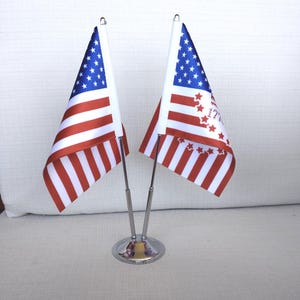 May include: Two small American flags with red and white stripes and a blue field of stars. The flags are displayed on a silver-colored stand, suitable for a desk or table. One flag has text and stars.