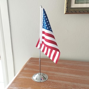 May include: A small American flag on a silver metal stand. The flag features red and white stripes and a blue field with white stars. The stand has a round base and a thin pole.